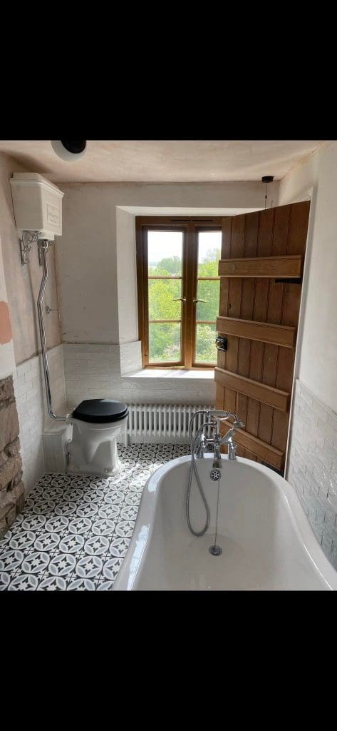 A rustic bathroom design features a clawfoot bathtub with a chrome faucet and a high flush toilet. A wooden door with a window opens to greenery, enhancing its charm. The floor showcases black and white patterned tiles, adding elegance to this luxury bathroom space.