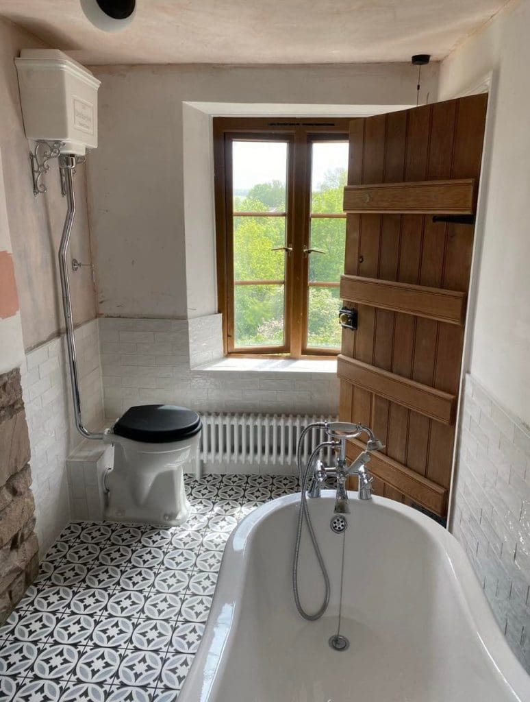 This luxury bathroom design boasts a vintage-style bathtub with a window view of lush greenery. The floor showcases black and white patterned tiles, complemented by a high cistern toilet with a black seat. A partially open wooden door and white brick tile walls add to its charm.