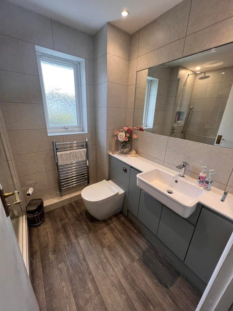 A modern bathroom design with grey tiles and wooden flooring features a large mirror, a sink with cabinets, a wall-mounted toilet, and a glass shower. A heated towel rack and a vase with flowers enhance the luxury feel near the frosted window.