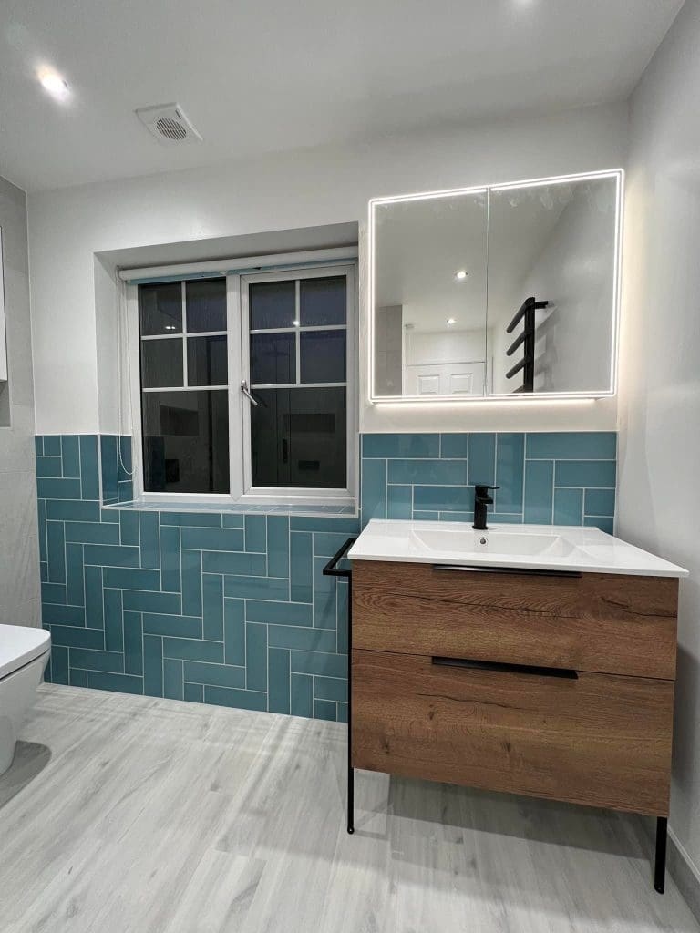 A luxurious bathroom installation showcases teal herringbone tiles, a wooden vanity with a sleek black faucet, and an illuminated mirror above. A grid-patterned window graces the tiled wall, while light wood-like tiles cover the floor, epitomizing modern bathroom design.