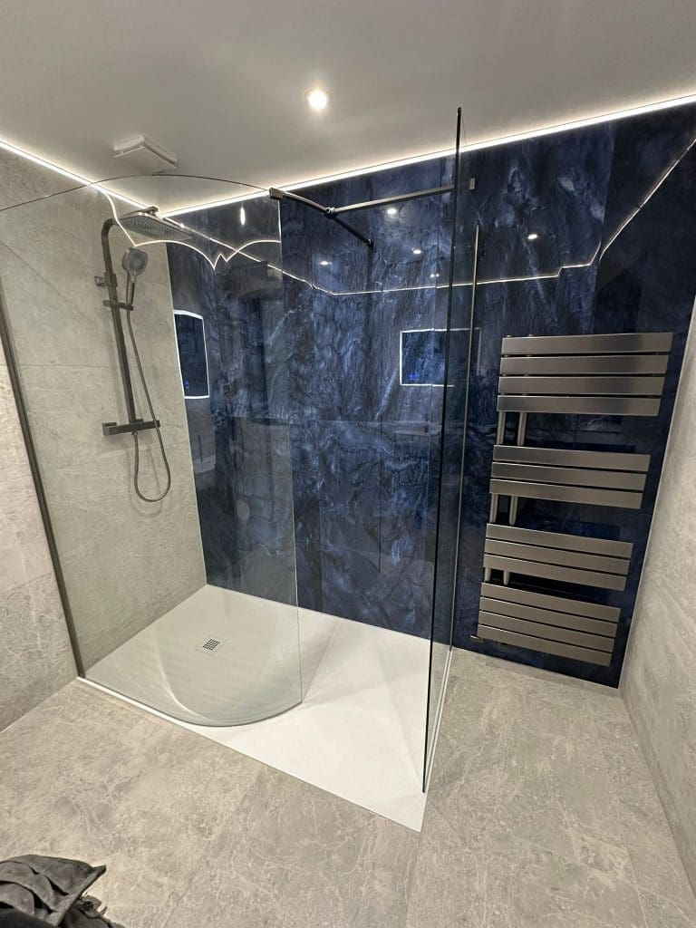Luxury bathrooms feature modern elements like a glass-enclosed shower with a black and dark blue textured wall. This bathroom design is complete with a rainfall showerhead, light grey tile flooring, and a sleek horizontal metal towel rack for an elegant touch.