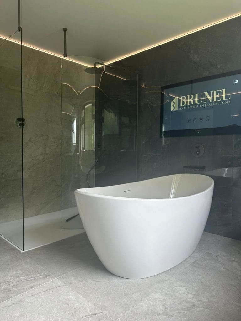 Modern bathroom featuring a freestanding white bathtub, a glass-enclosed shower, and a mounted screen displaying "Brunel Bathroom Installations" on the back wall. This luxury bathroom showcases sleek, minimalist design with gray stone tiles, exemplifying premium bathroom installations.