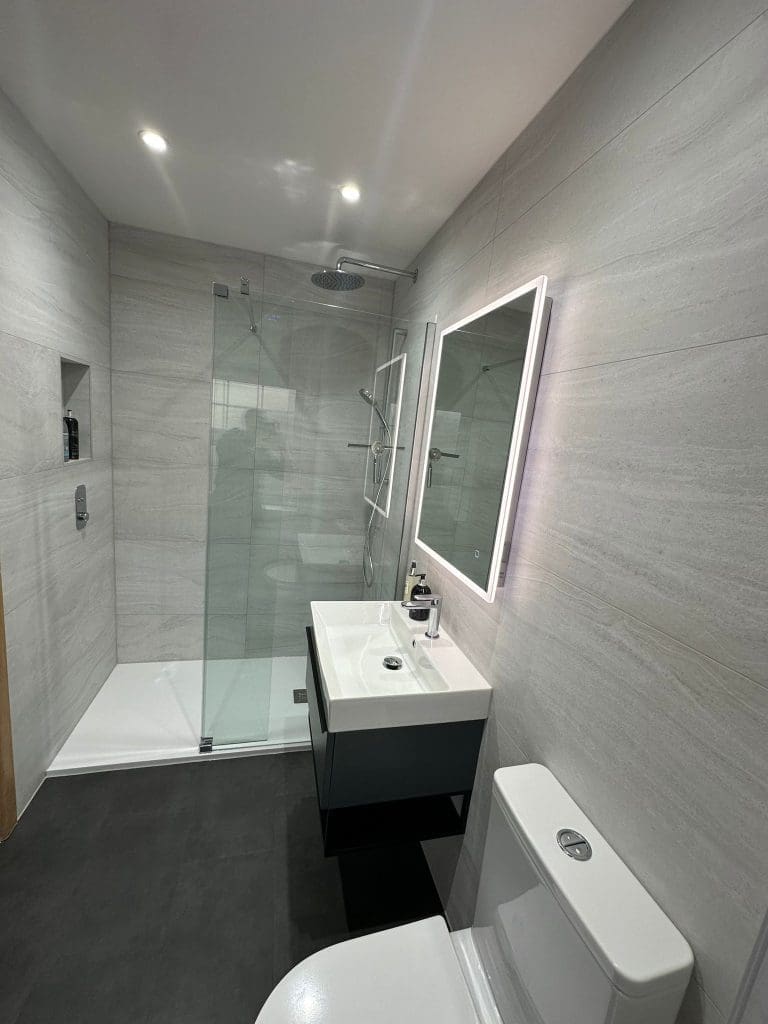 Experience bathroom luxury with a modern walk-in shower featuring a glass door, rainfall showerhead, and elegant grey wall tiles. The room boasts a white sink with an illuminated mirror above and a pristine toilet. The dark gray floor completes the sophisticated bathroom design.