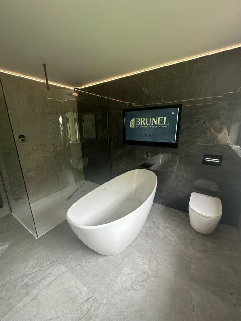 A showcase of luxury bathroom design, this modern space features a freestanding white bathtub, a glass-enclosed shower, and a wall-mounted toilet. The dark gray tiled walls and floor complement the elegance of the "BRUNEL" screen above the toilet.