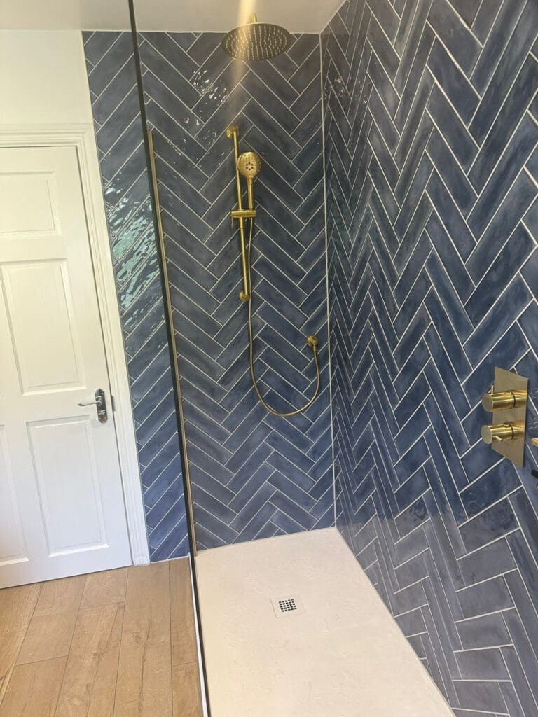 A modern luxury bathroom design features a shower with blue herringbone tiles and gold fixtures, including a rain shower head and handheld option. The white floor is sleek, contrasting beautifully with the glimpse of a wooden floor and white door just outside the shower area.