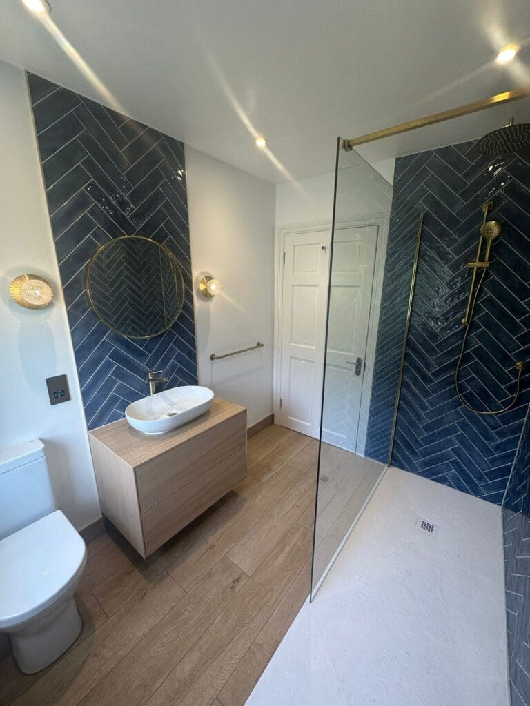 This luxury bathroom design boasts blue herringbone tile walls and includes refined bathroom installations like a glass-enclosed shower with a rain showerhead, a wooden vanity beneath a round mirror, and a sleek white sink. The floor is light wood, enhanced by soft, warm lighting for an exquisite touch.