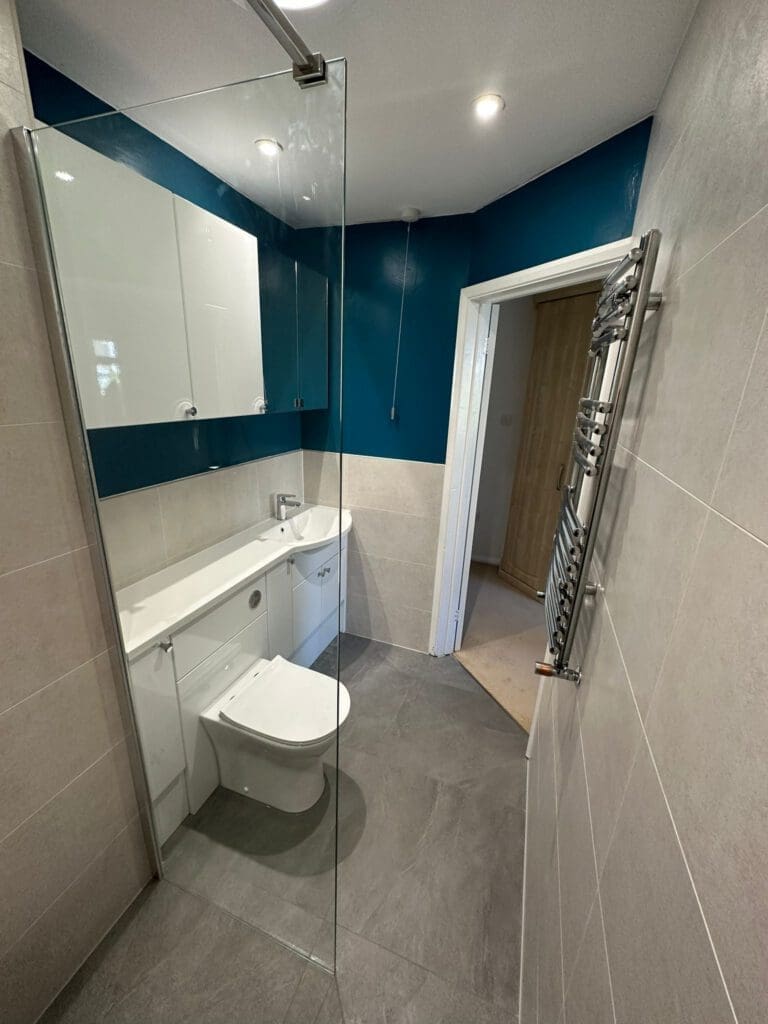 Experience luxury bathroom design in this modern space featuring a glass shower partition, wall-mounted white toilet, and sleek vanity. Teal and beige tiled walls complement recessed lighting, while a stainless steel heated towel rail adds elegance to the gray tile flooring.