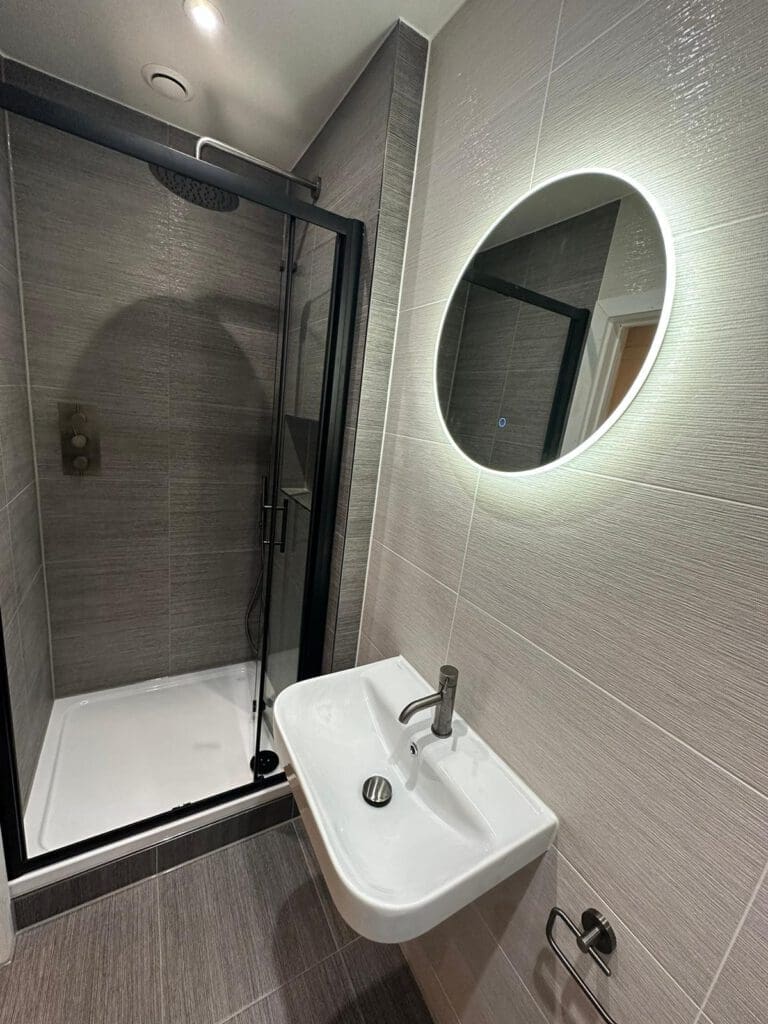 A modern bathroom design features a glass shower enclosure with a dark frame, a wall-mounted oval mirror with lighting, and a white rectangular sink with a silver faucet and soap dispenser. Sleek gray tiles cover the walls and floor, encapsulating the feel of luxury bathrooms.