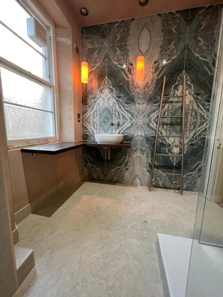 A modern bathroom design with marble-patterned wall tiles, a white wall-mounted sink, and a wooden shelf. An orange wall light illuminates the space. A wooden ladder leans against the wall. Large window on the left and a shower area on the right offer luxury in every detail.
