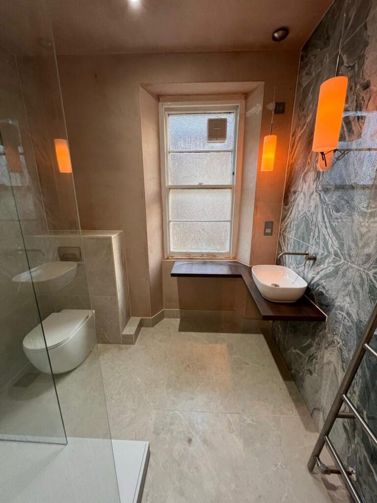 A modern luxury bathroom features a large frosted window, a wall-mounted sink on a wooden shelf, and a toilet. Marble tiles partly cover the walls. Two orange pendant lights offer warm lighting over light-colored stone flooring, showcasing exquisite bathroom design.