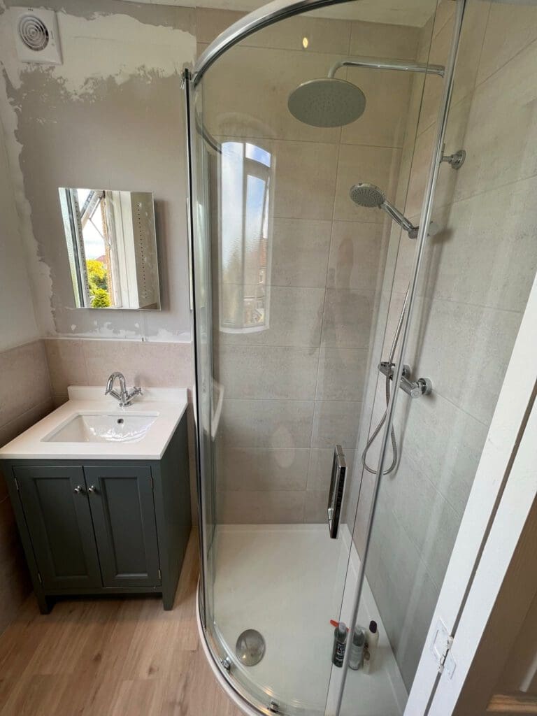 A modern bathroom exuding elegance with its glass-enclosed corner shower featuring a rainfall showerhead. Adjacent is a white sink atop a dark green vanity, complemented by a small mirror. This luxury bathroom showcases partial painted walls and light wood flooring for exquisite bathroom design.