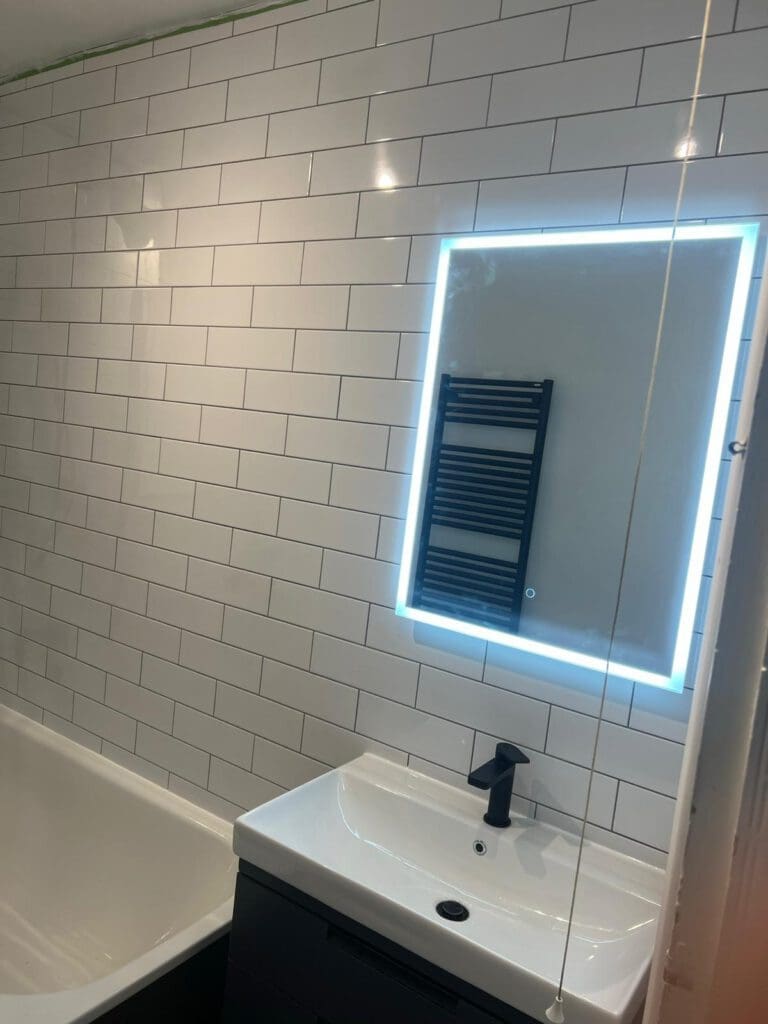 A modern bathroom design with white subway tiles, a sleek white bathtub on the left, and a chic white sink featuring a black faucet below an illuminated mirror. A black towel warmer adds a touch of elegance, embodying luxury bathrooms at their finest.