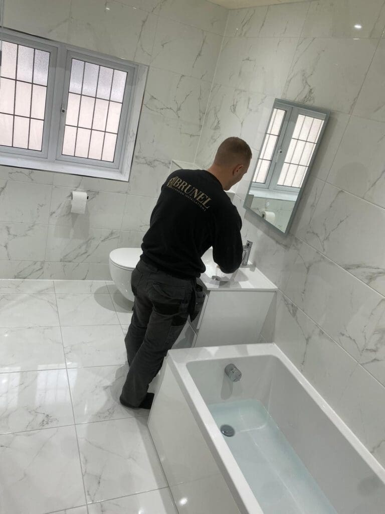 A man in a black sweatshirt stands in a modern white marble bathroom, working at a sink counter. The room, part of a luxury bathroom design, features a bathtub, a toilet, and a window with frosted glass.
