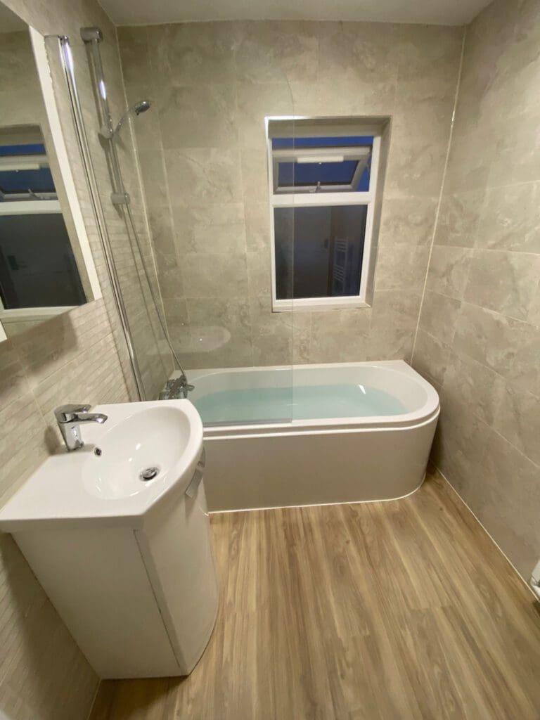 A modern bathroom design showcasing a bathtub with a glass shower screen, wall-mounted shower head, and white sink with sleek faucet. A mirror sits above it all, framed by neutral-toned tiles and wood-effect vinyl flooring, epitomizing luxury bathrooms at their finest.
