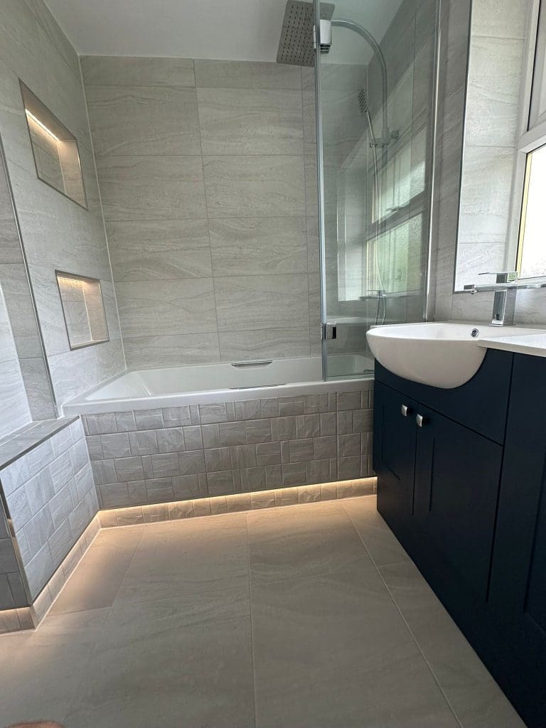 Discover a luxury bathroom design featuring modern elements like large gray tiles and a bathtub with built-in shelving. Under-cabinet lighting highlights the dark vanity, perfectly placed under a window to welcome natural light, creating an elegant and inviting space.