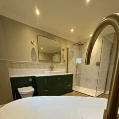A modern luxury bathroom featuring a large white bathtub, a green vanity with dual sinks, gold fixtures, a large mirror, and a glass shower enclosure with a rainfall showerhead. The walls are light with subtle paneling, and the warm lighting completes this exquisite bathroom design.