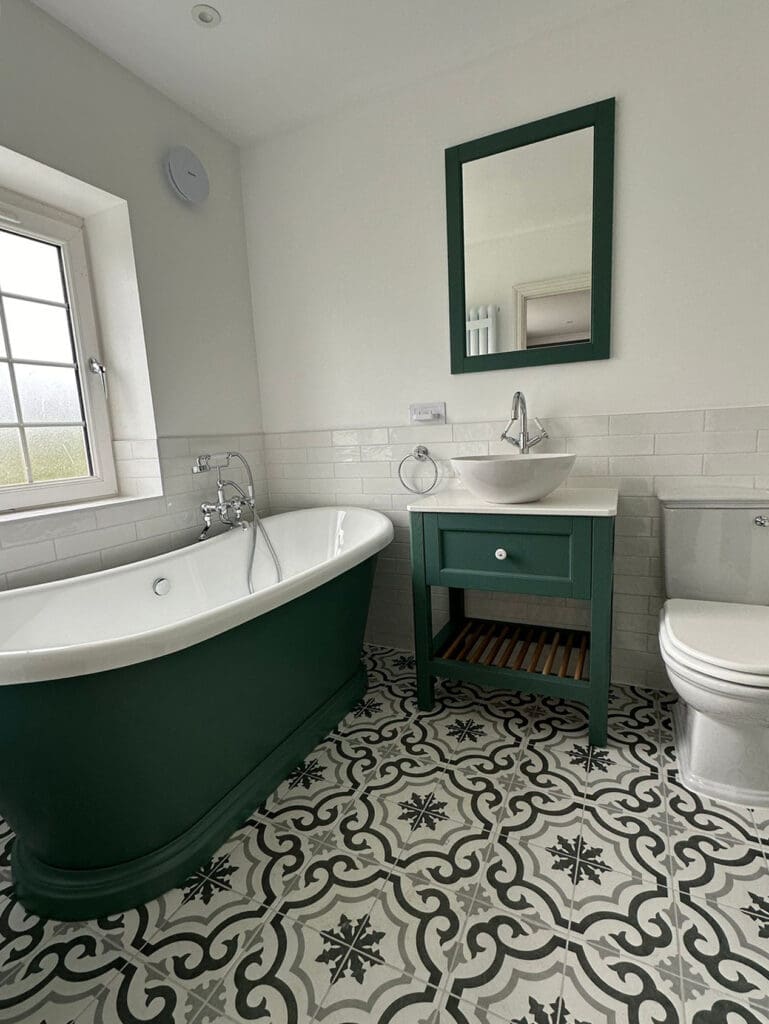 A bathroom design featuring a green freestanding bathtub and a matching vanity with a white sink and mirror above. The floor boasts black, white, and gray patterned tiles, while a window floods the space with natural light. This luxury bathroom exudes modern elegance.