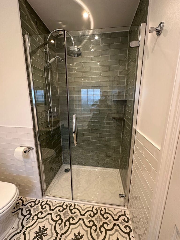 A modern bathroom showcases luxury bathroom design with a glass-enclosed shower adorned in dark green subway tiles. The floor displays a black and white intricate pattern, complemented by a chrome showerhead and handle, alongside a toilet and wall-mounted toilet paper holder.