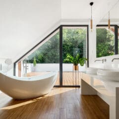 Modern bathroom with a sloped ceiling, featuring a sleek freestanding bathtub, dual sinks on a long countertop, and large windows overlooking a deck with greenery. The luxury bathroom is enhanced by warm wooden flooring and minimalistic decor that complete the sophisticated design.