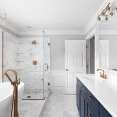 A modern luxury bathroom features white marble tile, a glass shower, freestanding tub, and a double sink vanity with blue cabinets. Gold fixtures accent the room, while a large window bathes it in natural light. A chandelier elegantly hangs from the ceiling.